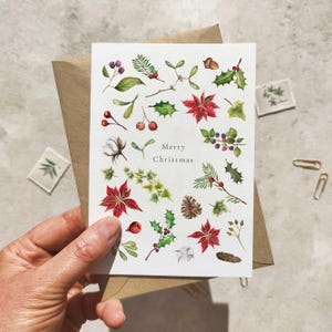 May include: A white greeting card with watercolor illustrations of Christmas foliage, including poinsettias, holly, and berries. The words "Merry Christmas" are printed on the card. The card is held in front of a brown envelope.