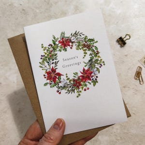 May include: A white greeting card with a watercolor wreath of red poinsettias, green leaves, and berries. The text "Season's Greetings" is centered within the wreath. The card is paired with a brown envelope.