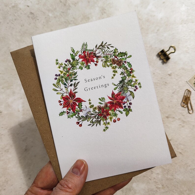 May include: A white greeting card with a watercolor wreath of red poinsettias, green leaves, and berries. The text "Season's Greetings" is centered within the wreath. The card is paired with a brown envelope.