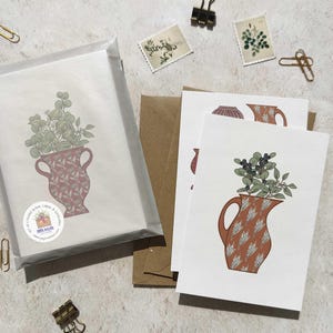 May include: Blank cards with a botanical illustration of a vase filled with greenery. The vase is a reddish-brown color with a leaf pattern. The cards are packaged in a clear cellophane sleeve with a sticker. Additional cards and envelopes are visible.