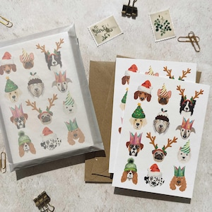 May include: A set of greeting cards featuring illustrated dog faces wearing festive hats and antlers. The dogs are depicted in various breeds and colors, with hats in red, green, and gold. The cards are packaged in a clear cellophane sleeve.