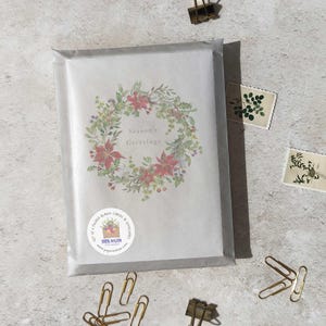 May include: A set of folded blank cards and envelopes in a clear cellophane package. The card features a wreath of red poinsettias and greenery with the text "Season's Greetings". Gold paper clips and stamps are scattered around the package.