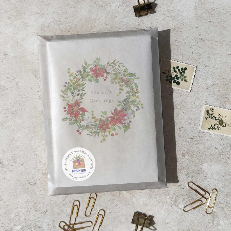 May include: A set of folded blank cards and envelopes in a clear cellophane package. The card features a wreath of red poinsettias and greenery with the text "Season's Greetings". Gold paper clips and stamps are scattered around the package.