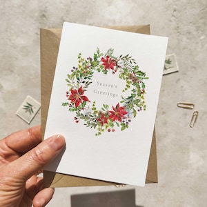 May include: A white greeting card with a watercolor wreath design featuring red poinsettias, green leaves, and berries. The card reads "Season's Greetings" and is held over a brown envelope.