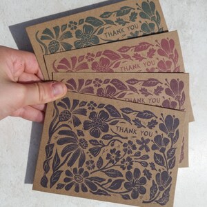 May include: A stack of four rectangular kraft paper cards, each featuring a unique floral print in a different ink color and the text THANK YOU printed in a bold, sans-serif font. The botanical illustrations cover the entire card surface.