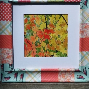 May include: A white picture frame with a patchwork design border. The frame is decorated with colorful fabric squares featuring birds, flowers, and geometric patterns. The frame contains a photograph of a tree with orange and yellow leaves.