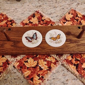 May include: A wooden wall-mounted coat rack with two pegs and two round ceramic coasters. The coasters feature butterfly illustrations in shades of blue, orange, and brown. The rack is made of dark wood.