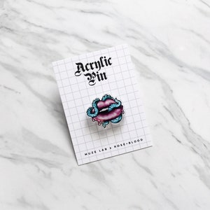 May include: A blue and pink acrylic pin featuring a pair of lips with a snake wrapped around them. The pin says "Acrylic Pin" and "MUZE LAB x ROSE-BLOOD".
