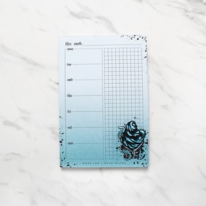 May include: A blue and white weekly planner notepad with a grid on the right side and a space for daily to-do lists on the left. The notepad has a black and blue graphic of a rose with a splatter effect around it. The bottom of the notepad says "MUTE LAB x ROSEBLOOD".