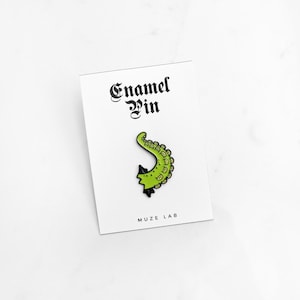 May include: A green and black enamel pin in the shape of a tentacle, displayed on a white card. The card reads "Enamel Pin" in a gothic font, with "MUZE LAB" printed below. The tentacle pin has a detailed design.