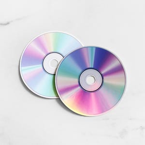 May include: Two circular compact discs featuring a reflective, iridescent surface that displays a spectrum of rainbow colors. Each disc has a central hole and a smooth, metallic finish that catches the light, creating shifting gradients of blue, pink, and green.