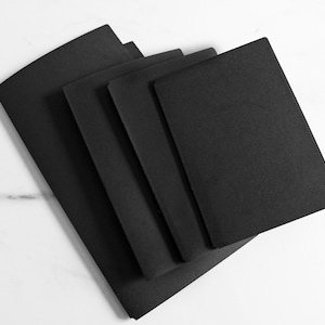 May include: A set of four rectangular black notebooks arranged in a staggered, overlapping stack. Each notebook features a solid matte black cover with a clean, simple design and rounded corners, presented in a uniform vertical orientation.