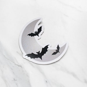 May include: A silver glitter crescent moon sticker with three black bats flying around it.
