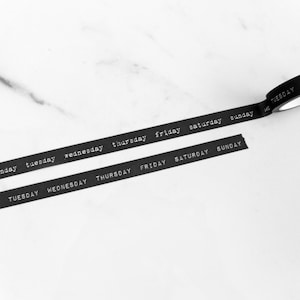 May include: Black washi tape with the days of the week printed in white. The tape is unrolled, with two cut pieces and a roll visible. The days of the week are printed in a simple sans-serif font.