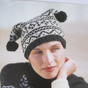 May include: A black and white knit hat with a pointed top and two black pom-poms. The hat features a geometric pattern and is worn with a matching sweater. The sweater has a black background with white geometric designs.