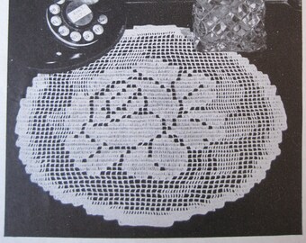 May include: A white crocheted doily with a delicate floral design. The doily is laid out on a dark surface with a vintage rotary phone and a glass vase of flowers in the background.
