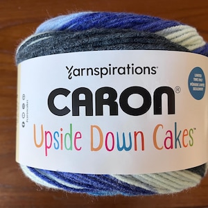 May include: A ball of Caron Upside Down Cakes yarn featuring a white paper label with the text Yarnspirations, CARON, and Upside Down Cakes. The yarn displays a variegated color pattern consisting of dark charcoal gray, royal blue, and pale mint green.