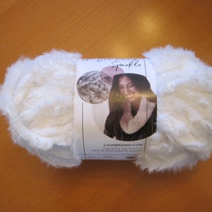 May include: A skein of white faux fur yarn with silver sparkle threads. The yarn is labeled "Lion Brand Sparkle" and has a photo of a woman wearing a white faux fur scarf.