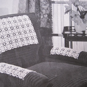 Op de afbeelding: Een close-up van een gehaakte stoelset met witte doilies op de rugleuning en armleuningen van een donkergrijze fauteuil.