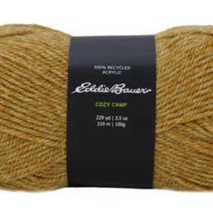 Puede incluir: Un ovillo de hilo Eddie Bauer Cozy Camp en un cálido color marrón dorado. La etiqueta dice "100% acrílico reciclado" y "Cozy Camp" con el metraje y el peso indicados como "229 yd | 3.5 oz | 210 m | 100g".