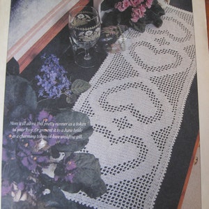 May include: White crocheted table runner with a delicate floral pattern. The runner is laid on a dark surface with purple flowers in the foreground. The text "June 2000 Crochet World 35" is visible in the bottom right corner.