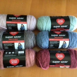 May include: Six skeins of Red Heart Super Saver Brushed yarn in shades of pink, grey, mint green, blue, and burgundy. Each skein is labelled with the brand name and product details. Each skein is 141g.