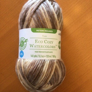 Könnte beinhalten: Eine Knäuel Eco Cozy Watercolors Garn in einer braunen, beige und weißen Farbkombination. Das Garn besteht aus recyceltem Kunststoff und ist 145 Yards / 5,3 Unzen oder 133 Meter / 150 Gramm. Das Etikett gibt auch an, dass ein Muster enthalten ist.
