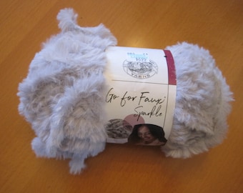 May include: A skein of light gray faux fur yarn with a label that reads "Go for Faux Sparkle." The yarn is fluffy and soft, ideal for knitting or crocheting projects. The label also features a picture of a woman.