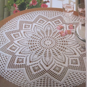 May include: A white crocheted tablecloth with a delicate floral pattern. The tablecloth is laid on a brown wooden table with two glasses of water and a few small boxes of sweets on the table.