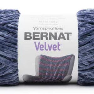 May include: A skein of Bernat Velvet yarn in a dusty blue color. The label displays the brand name and a picture of a knitted blanket. The yarn is soft and plush, ideal for knitting and crochet projects.