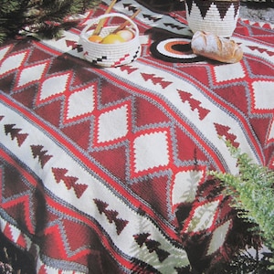 May include: A picnic scene featuring a red, white, and gray patterned blanket. Two woven baskets, one with fruit and wooden utensils, the other with a pineapple. A loaf of bread and a coaster are also on the blanket.