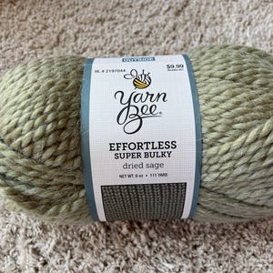 May include: A skein of Yarn Bee Effortless super bulky yarn in the color dried sage. The white paper label features a bee icon, the text Yarn Bee, Effortless Super Bulky, dried sage, and states a net weight of 6 oz and length of 111 yards.