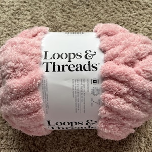 May include: A skein of soft, light pink jumbo yarn with a white paper label featuring the text Loops & Threads. The label indicates 100% polyester material, hand wash instructions, and a size 7 jumbo weight classification for this craft supply.