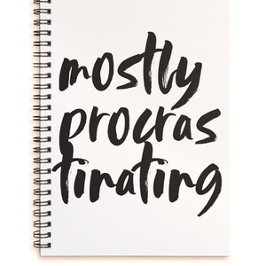 Pode incluir: Um caderno branco com espiral com as palavras "mostly procrastinating" escritas em preto com pincel. O caderno está aberto e o texto está centrado na página.