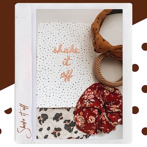 Puede incluir: Una libreta blanca con lunares negros y el texto "shake it off" escrito en naranja. La libreta está rodeada de una diadema de estampado de leopardo marrón y blanco, una scrunchie floral roja y una diadema tejida marrón.