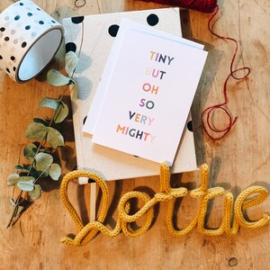 May include: A selection of stationery items on a wooden surface. Includes a roll of polka dot washi tape, a notebook with black polka dots, a greeting card with the text "TINY BUT OH SO VERY MIGHTY", a yellow knitted word "Lottie", and red twine.