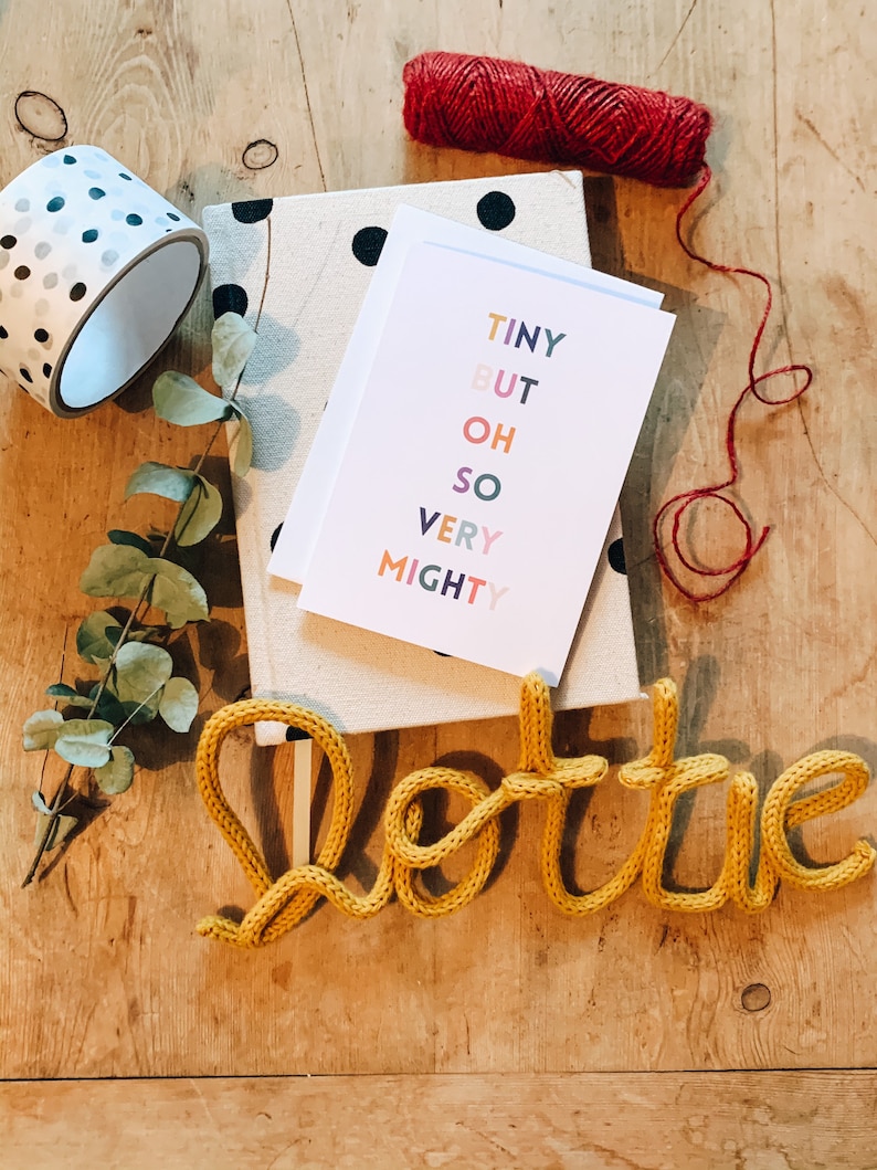 May include: A selection of stationery items on a wooden surface. Includes a roll of polka dot washi tape, a notebook with black polka dots, a greeting card with the text "TINY BUT OH SO VERY MIGHTY", a yellow knitted word "Lottie", and red twine.