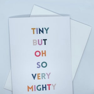 May include: A white greeting card featuring the phrase "TINY BUT OH SO VERY MIGHTY" in colourful block letters. The card is accompanied by a white envelope, ideal for sending messages.