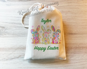 May include: A white canvas gift bag with a drawstring closure. The bag features three colorful Easter bunny illustrations and the words "Taylor" and "Happy Easter" in green script. The bag is from Kpont Gift Company.