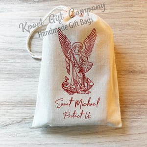 May include: A white drawstring bag with a red image of Saint Michael the Archangel, holding a spear and standing over a dragon. The bag has the text "Saint Michael Protect Us" printed on it.