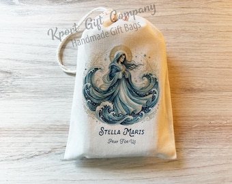 May include: A cream-colored drawstring gift bag featuring a printed illustration of a figure in a blue robe surrounded by stylized ocean waves and a golden halo. The text Stella Maris and Pray For Us is printed in blue below the central image.