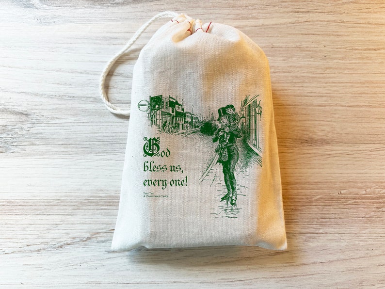 K&ouml;nnte beinhalten: Ein cremefarbener Baumwollbeutel mit Kordelzug und einem gr&uuml;nen Druck, der eine Person zeigt, die ein Kind auf den Schultern durch eine viktorianische Stra&szlig;e tr&auml;gt. Der Text lautet: God bless us, every one! Tiny Tim, A Christmas Carol.