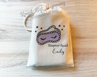 May include: A white drawstring gift bag with a sleep mask design in black and lavender, surrounded by black stars. The text "Sleepover Squad Emily" is printed below the mask. The bag is made of a canvas-like material.