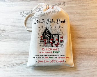 May include: A cream-colored drawstring canvas bag featuring a North Pole Bank graphic with elves and Santa Claus. The text reads: North Pole Bank, To: Jason Jones, For the Amount of $50.00, Christmas bank notes are legal tender, to be used for toys, candy, and all things fun! Santa Claus NPB Certified.