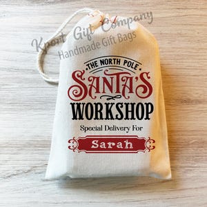 May include: White canvas gift bag with a red and black design that says "The North Pole. Santa's Workshop. Special Delivery For Sarah".