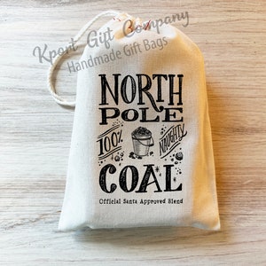 May include: A white drawstring bag with black text that reads "North Pole 100% Naughty Coal" with a small bucket of coal and a star. The bag is on a light wood surface.