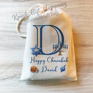 May include: A white fabric gift bag with a blue letter D, a menorah, a Star of David, and the text "Happy Chanukah David".
