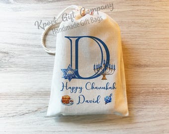 May include: A cream-colored drawstring gift bag featuring a large blue letter D, a Star of David, a menorah, and the text Happy Chanukah David. The design includes a small illustration of latkes and a dreidel in blue and gold tones.