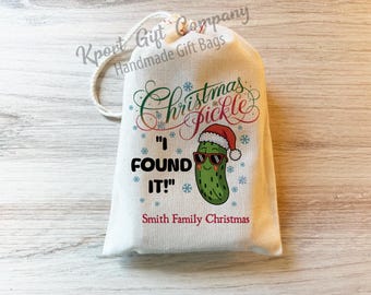 May include: A cream-colored drawstring gift bag featuring a green pickle wearing a red Santa hat and sunglasses. The text reads Christmas Pickle, I FOUND IT!, and Smith Family Christmas, surrounded by small blue snowflakes on the front.