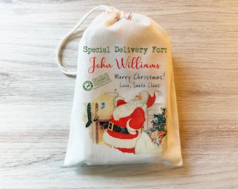 May include: A cream-colored drawstring gift bag featuring a printed illustration of Santa Claus near a fireplace. The text reads Special Delivery For: John Williams, Merry Christmas! Love, Santa Claus, and includes a 2028 postmark stamp graphic.